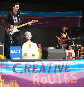 Creative Routes perform on stage. Beneath one of the electric guitars is a hospital ECT sign. There is a head covered in electrical wires connected to a huge plug.