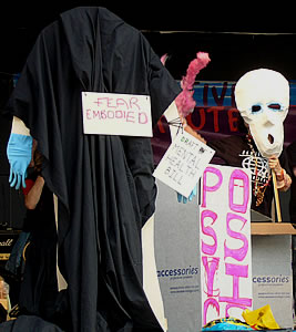 Some of the props on the stage. A black cloak carries the signs "fear embodied" and "draft mental health bill". A box is labelled with the word "psychosis". One of the performers holds a skull-shaped mask in front of his face.