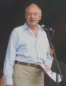 Ken Livingstone speaks into a microphone on the stage. In his left hand is his speech, while in his right hand (out of the picture) is a child he brought on stage with him