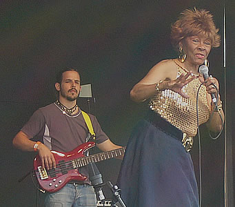 Totlyn Jackson giving a powerful singing performance. She looks like a true diva, wearing a shiny golden top and big earrings. Behind her is a member of her band, playing an electric guitar